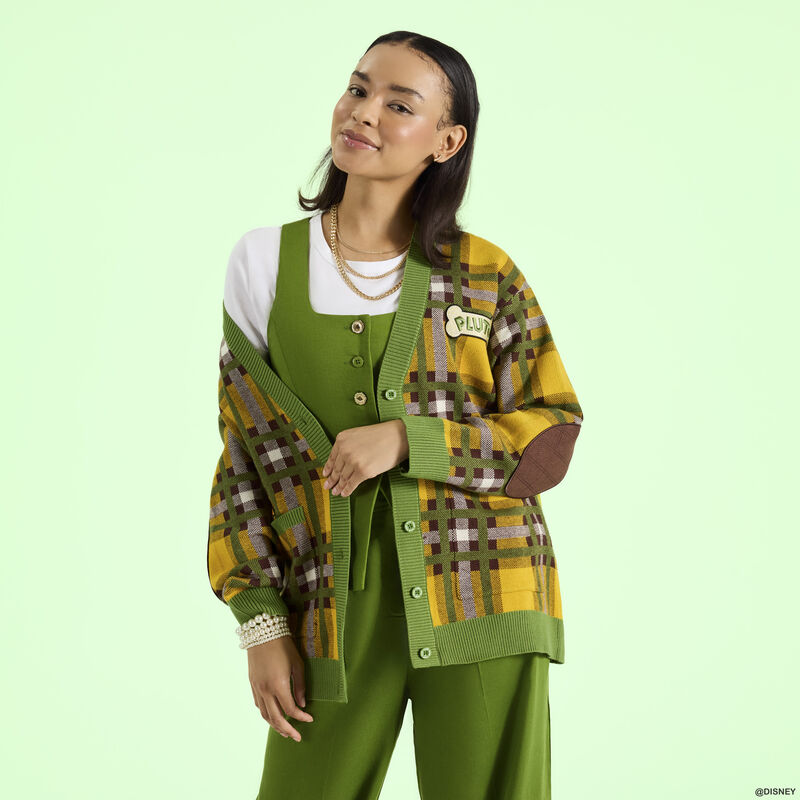 Woman wearing a green vest over a white top with green trousers. She wears the Loungefly Pluto 95th anniversary knit cardigan over the outfit. She stands in front of a green background.