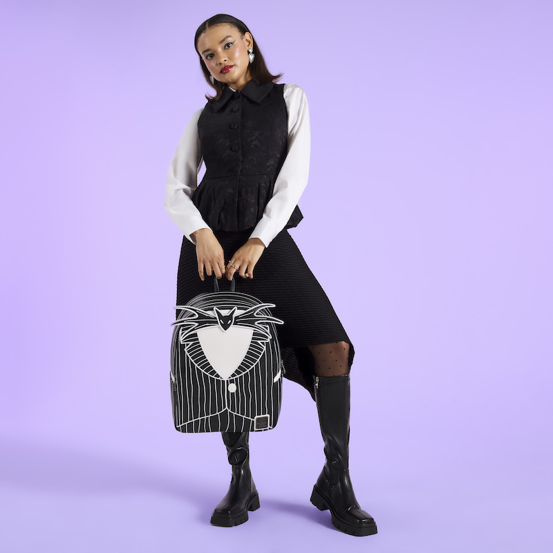 Woman wearing a black vest over a white long-sleeve top and a black skirt with black knee-high boots. She poses with the Loungefly Jack Skellington Full-Size Backpack. She holds it by the top handle. She stands in front of a purple background.