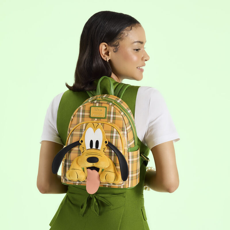 Woman facing away from camera wearing a green vest over a white top, she wears the Loungefly Pluto 95th Anniversary mini backpack on her back and stands in front of a green background.