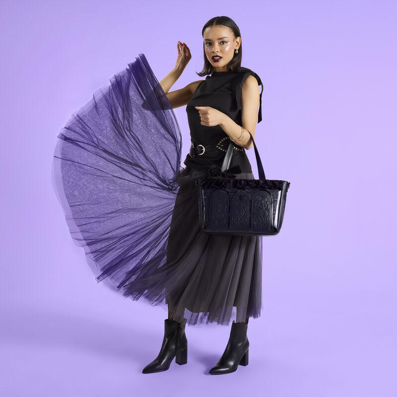 Woman wearing a black top, belt, and a tulle skirt with black boots. She wears the Loungefly Disney Villains Stained Glass Tote bag over an arm as she tosses the first layer of her skirt up in the air. She stands against a purple background.